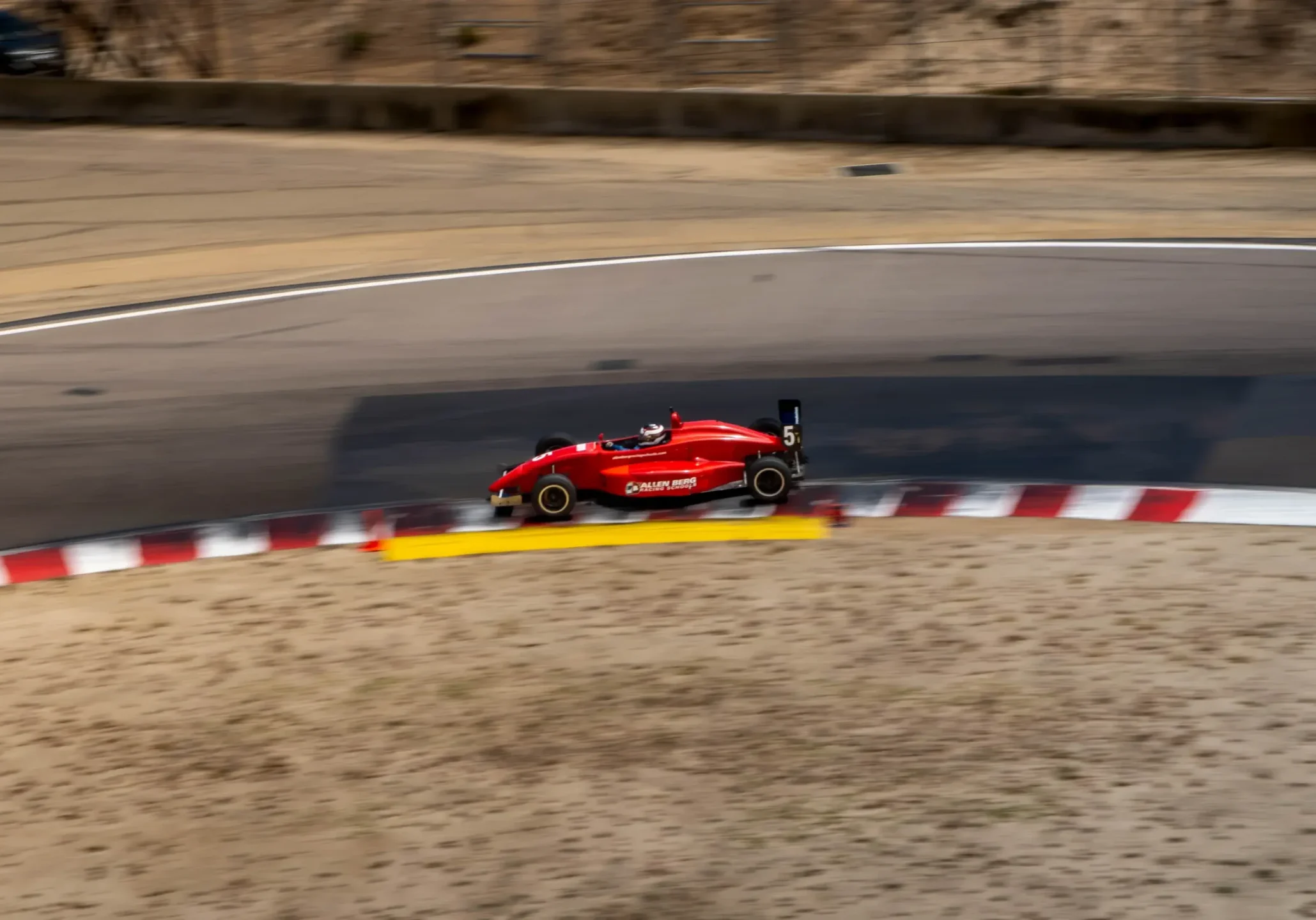 High-angle shot of red open-wheel race car number 5 driving near the red and white curbing on a race track