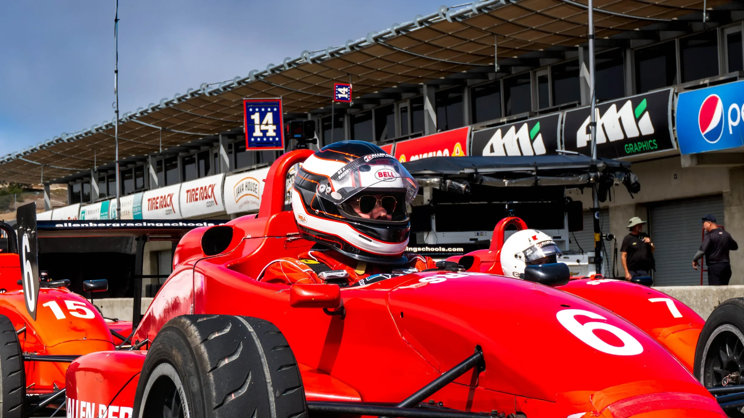 Close-up of a driver in a bell helmet seated inside red formula race car number 6 in the pit lane