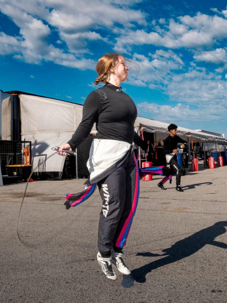 Another Podium for Berg Racing as Young Drivers Continue Strong Development at Road America 2 Female racing driver Ella White jumps rope during a pre-race warmup routine in the track paddock, wearing a black top and custom racing suit with her last name printed on the leg.
