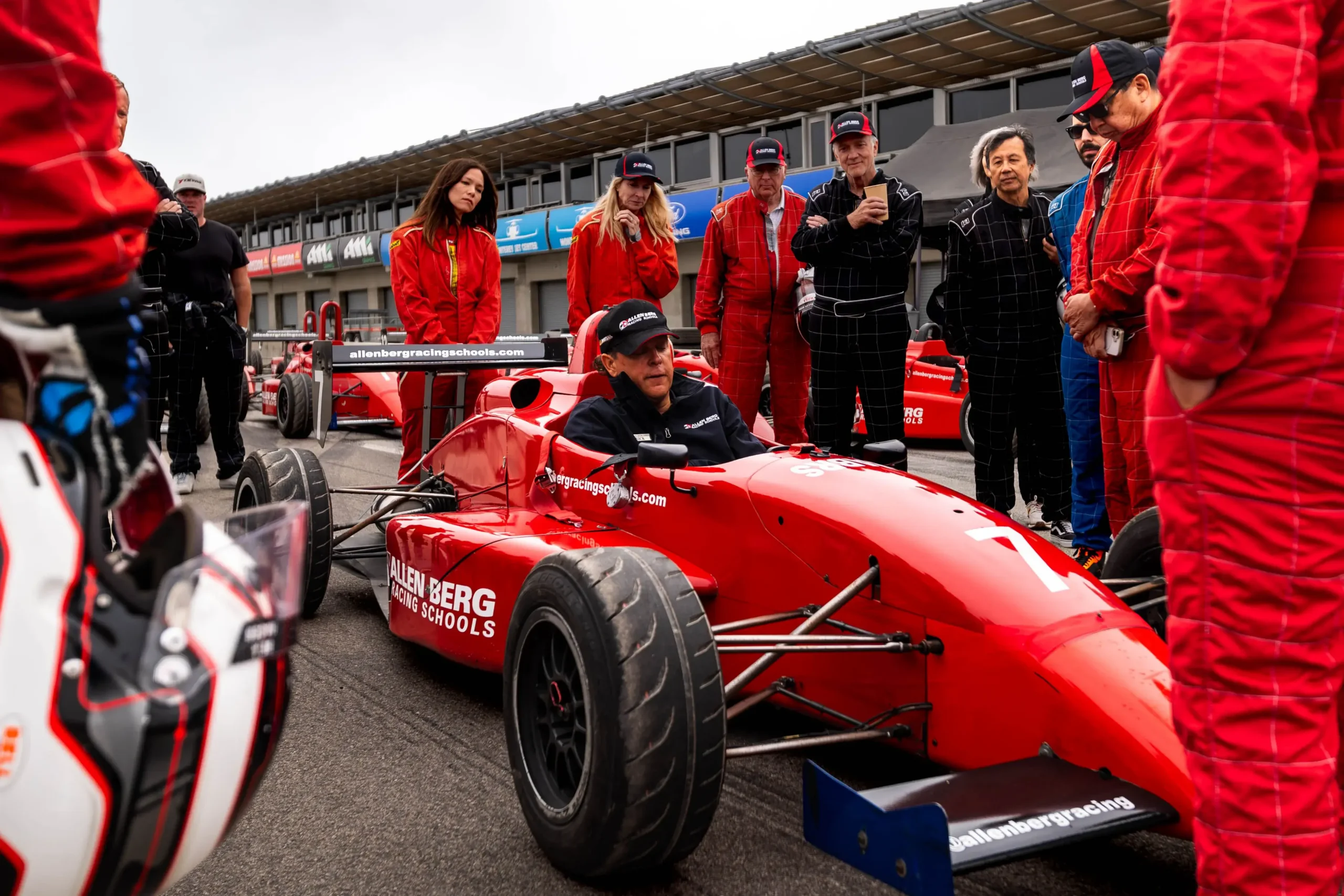 Instructor sitting inside a red formula race car explaining techniques to surrounding students in race suits