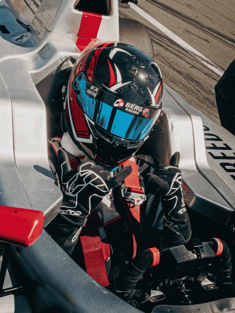 Another Podium for Berg Racing as Young Drivers Continue Strong Development at Road America 1 Formula racer Tyler Wade wearing a black and red Berg Racing helmet with a blue reflective visor, sitting in the cockpit of his silver race car while flashing a shaka hand gesture