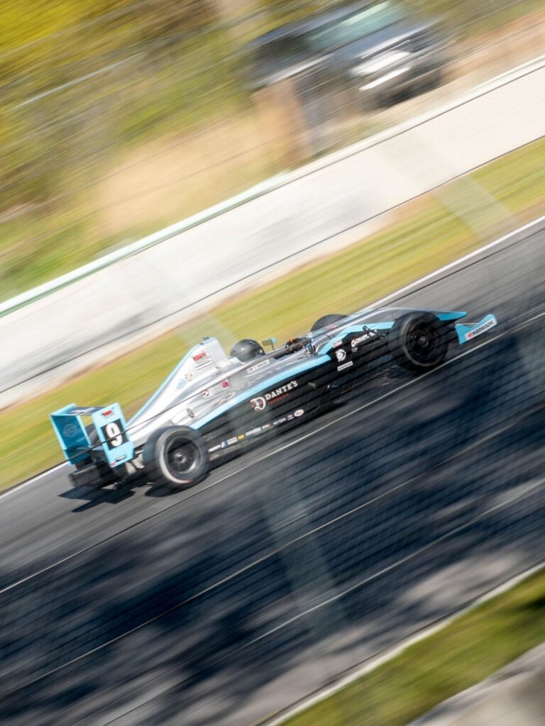 Another Podium for Berg Racing as Young Drivers Continue Strong Development at Road America 4 Tyler Wade's sleek race car speeds along a track, its blurred motion emphasizing velocity. The car is silver and teal, marked with sponsorship logos, under a sunny sky.